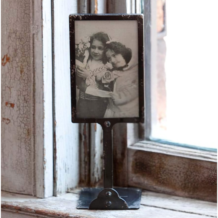 A vintage-style black metal tabletop photo frame with a distressed finish, holding a black-and-white photograph of two young girls, set on a rustic wooden windowsill.
