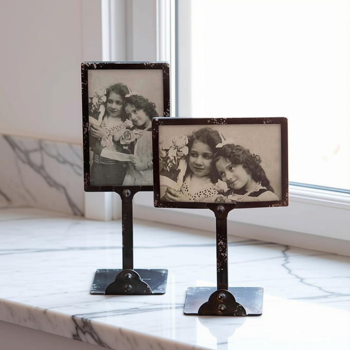Two vintage-style rectangular photo frames on metal stands displayed on a white marble windowsill. The frames hold sepia-toned black-and-white photographs of two young girls.