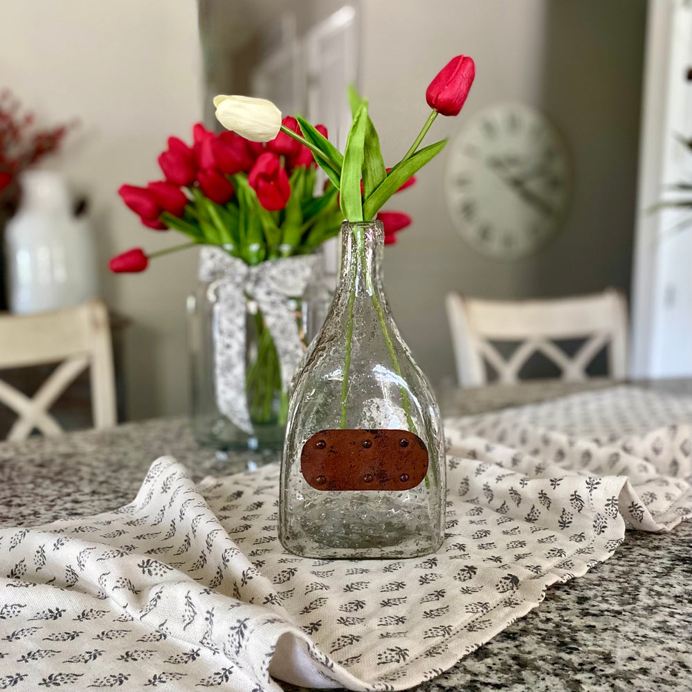 A clear glass bottle with a metal tag serving as a vase, filled with red tulips, placed on a table with a floral patterned cloth.