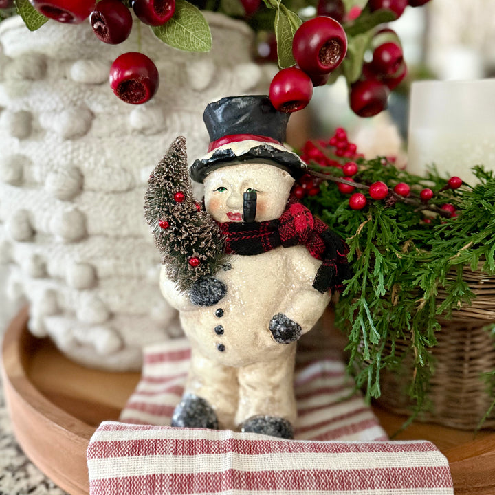 Decorative snowman figurine with a tree, surrounded by red berries and greenery on a striped surface.