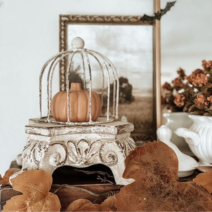 A small terracotta pumpkin displayed inside a distressed white birdcage atop an ornate vintage style pedestal riser, styled with dried leaves and autumn decor in a home setting.