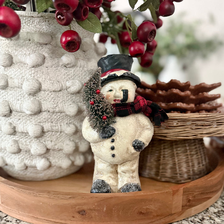 Decorative snowman figurine with a top hat and scarf on a wooden surface with berries and a textured vase in the background.