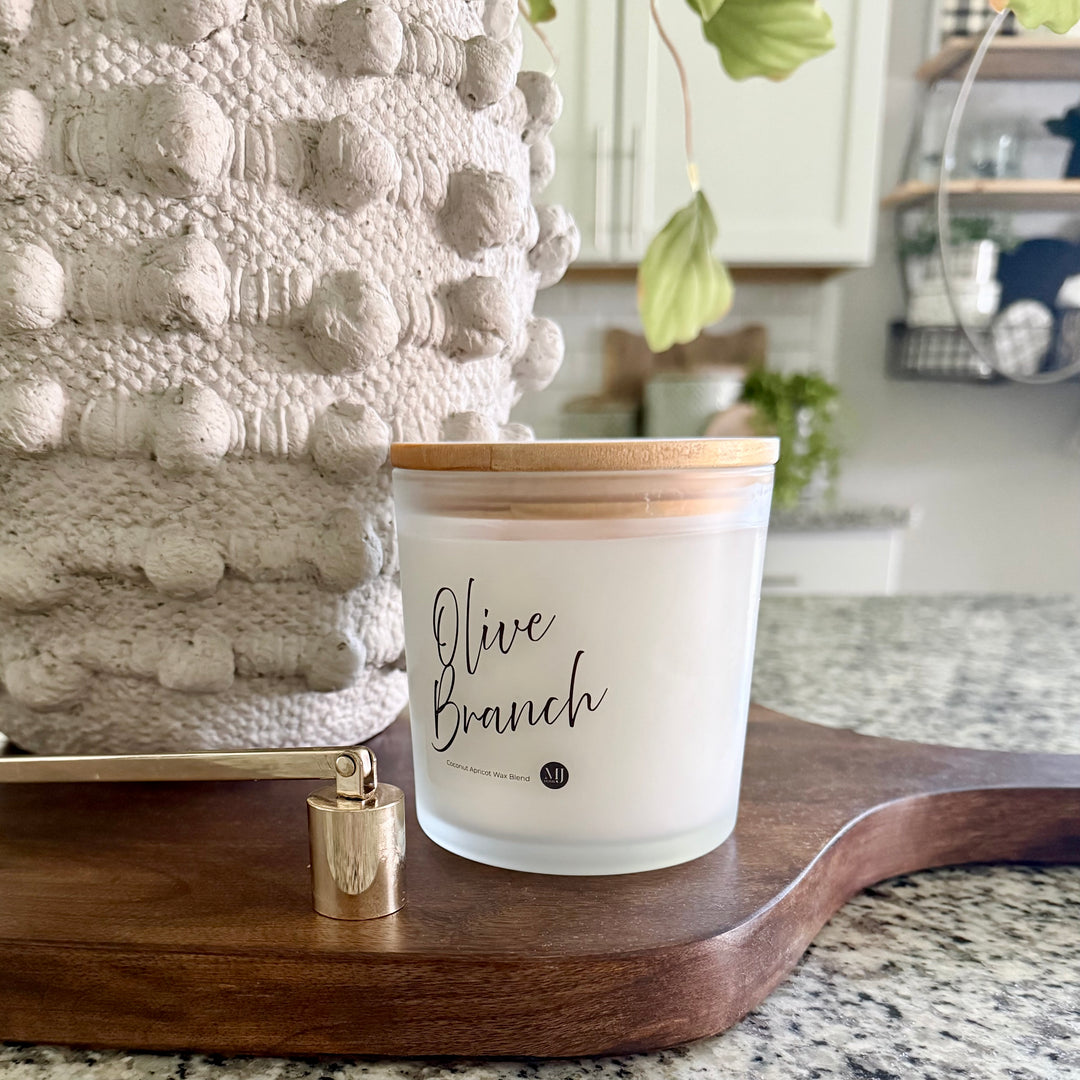 A frosted glass jar candle labeled 'Olive Branch' with a bamboo lid, styled on a mango wooden cutting board next to a brass candle snuffer in a kitchen setting.