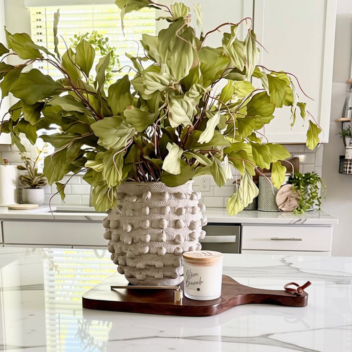 A large green leafy plant arrangement in a textured beige ceramic pot, displayed on a mango wood paddle style cutting board on a granite kitchen countertop.