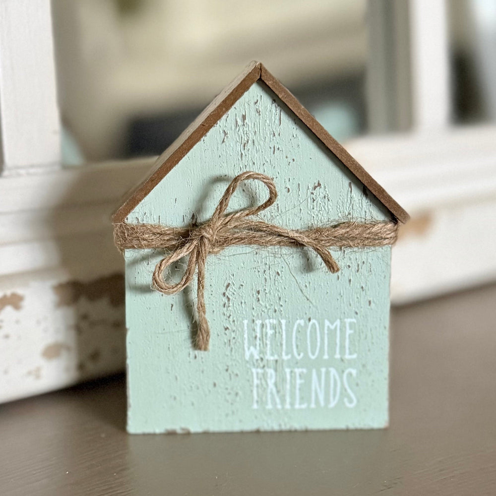 Small wooden house-shaped sign with 'Welcome Friends' text and a bow on a surface.
