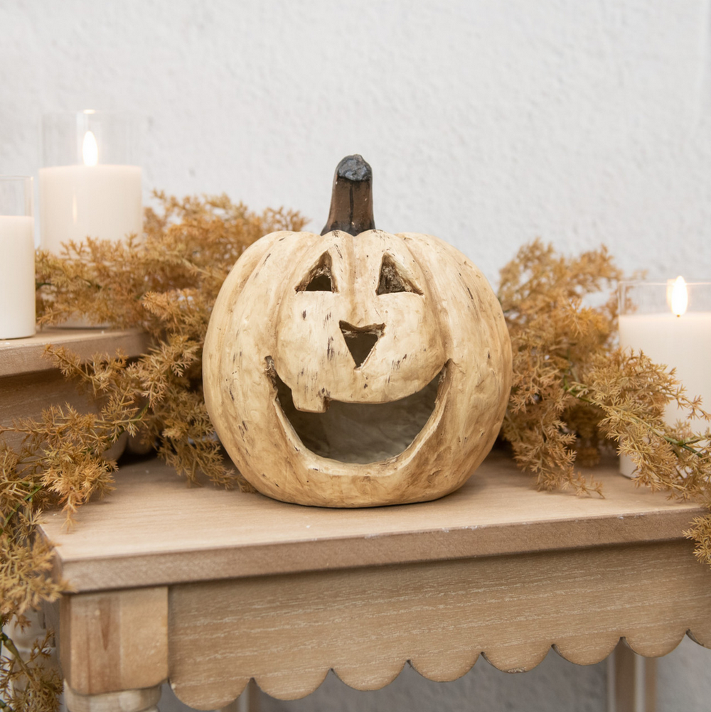 Decorative paper maché pumpkin with a carved face on a mantelpiece with candles and dried foliage.