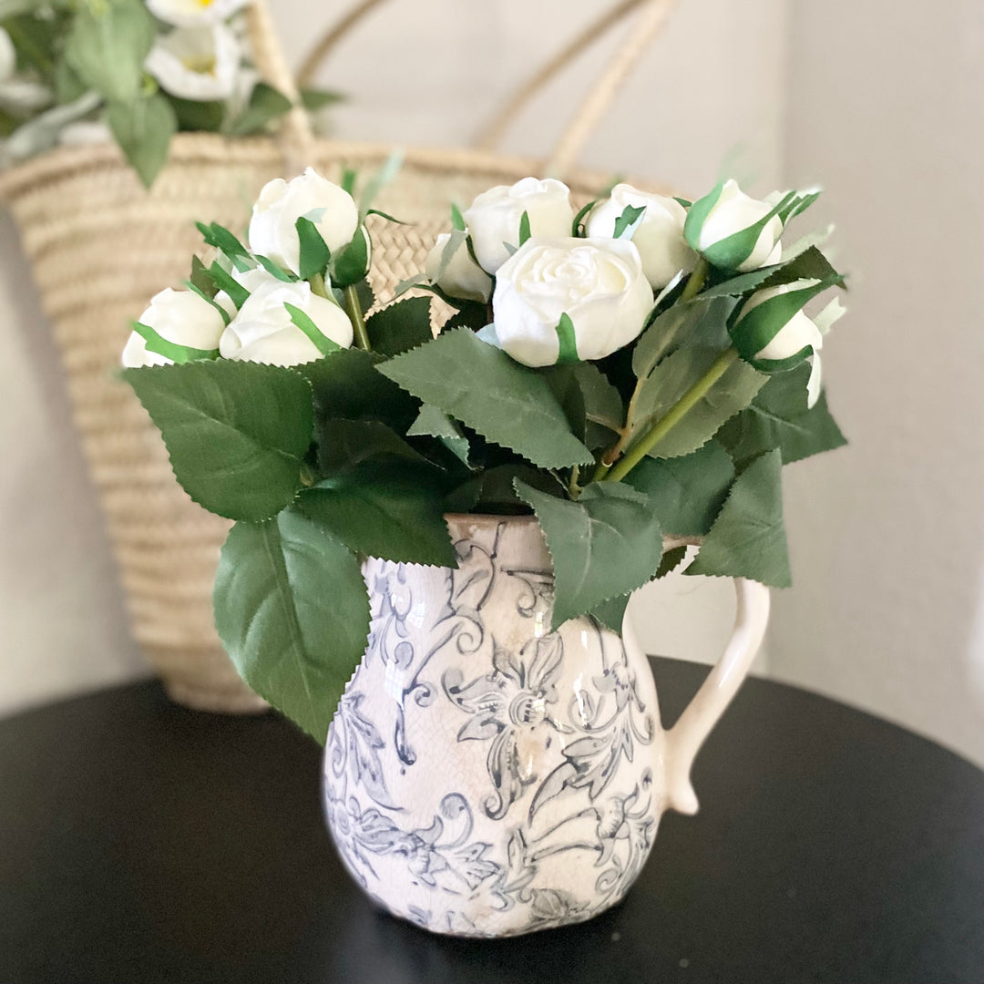 A bundle of artificial white cabbage roses with green leaves, tied together and placed in a patterned ceramic jug.