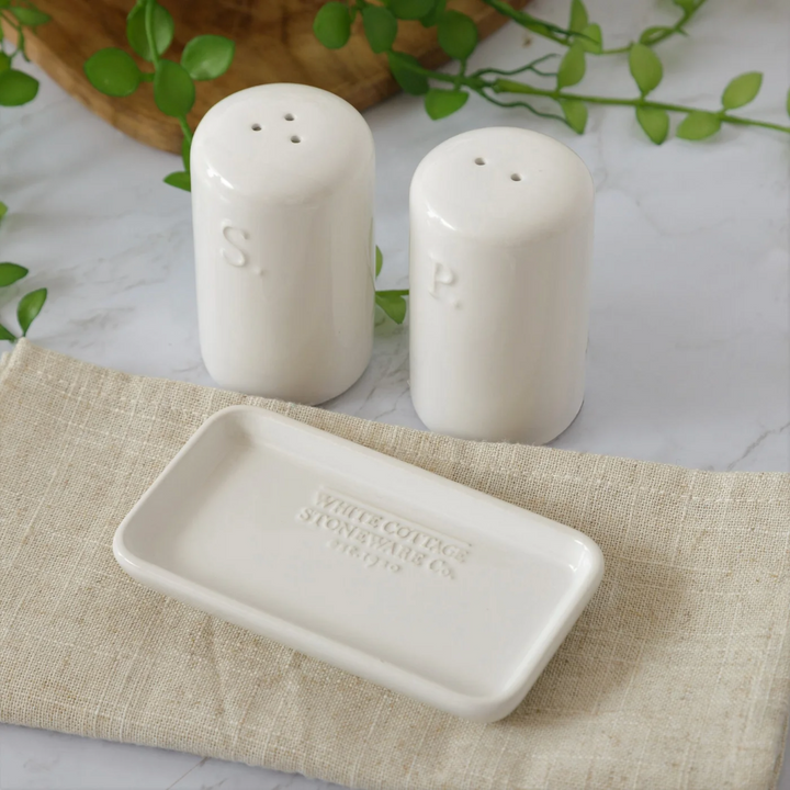 A white ceramic salt and pepper shaker set with embossed lettering, placed on a matching tray, presented on a table with a cloth and greenery in the background.