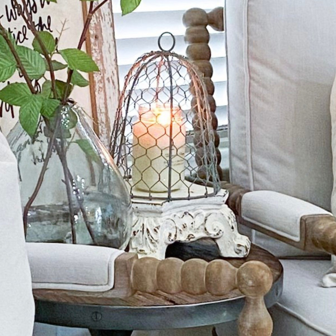 A decorative candle on top of an ornate white carved pedestal riser under a wire birdcage, styled on a wooden table between two upholstered chairs with a glass vase and greenery nearby.