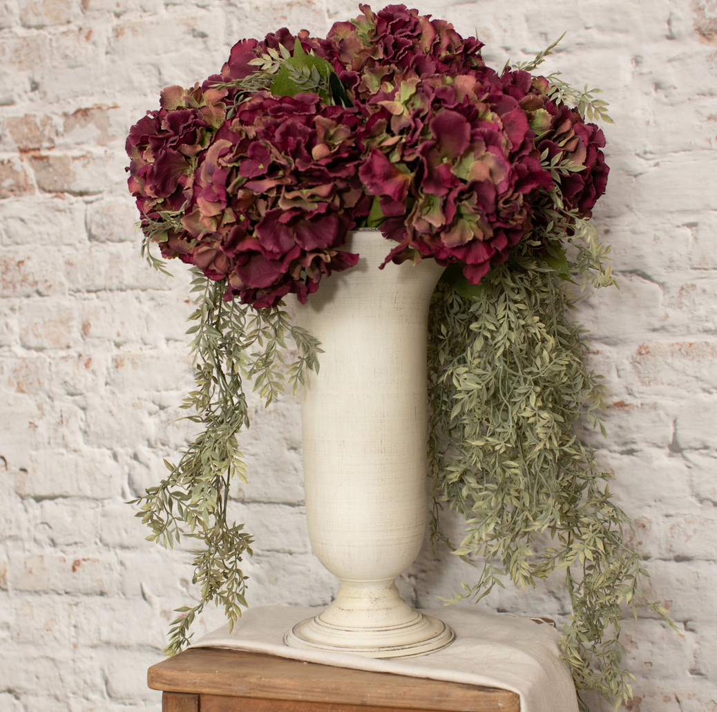 A fluted white vase with a distressed finish, filled with artificial flowers and greenery, placed on a surface against a brick wall backdrop.