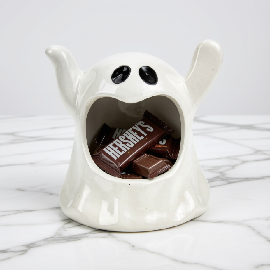 A white ceramic ghost-shaped candy holder filled with chocolate bars, including a Hershey's candy, set on a white marble surface against a light gray background.