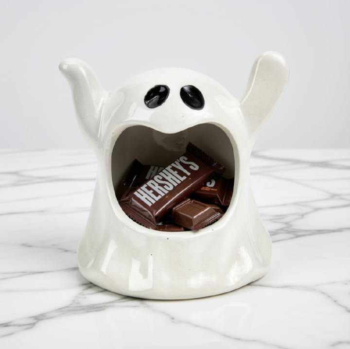 A white ceramic ghost-shaped candy holder filled with chocolate bars, including a Hershey's candy, set on a white marble surface against a light gray background.