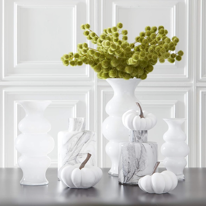Decorative arrangement with white vases, pumpkins, and a tall white vase with green foliage on a table.