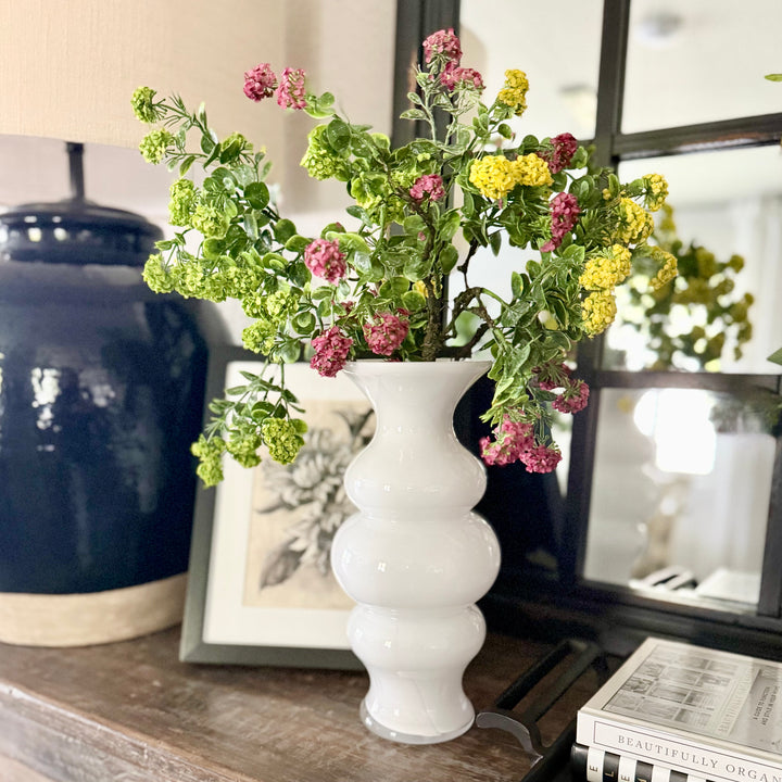 White vase with green, yellow and pink artificial blooming flowers on a wooden surface with decorative items in the background.