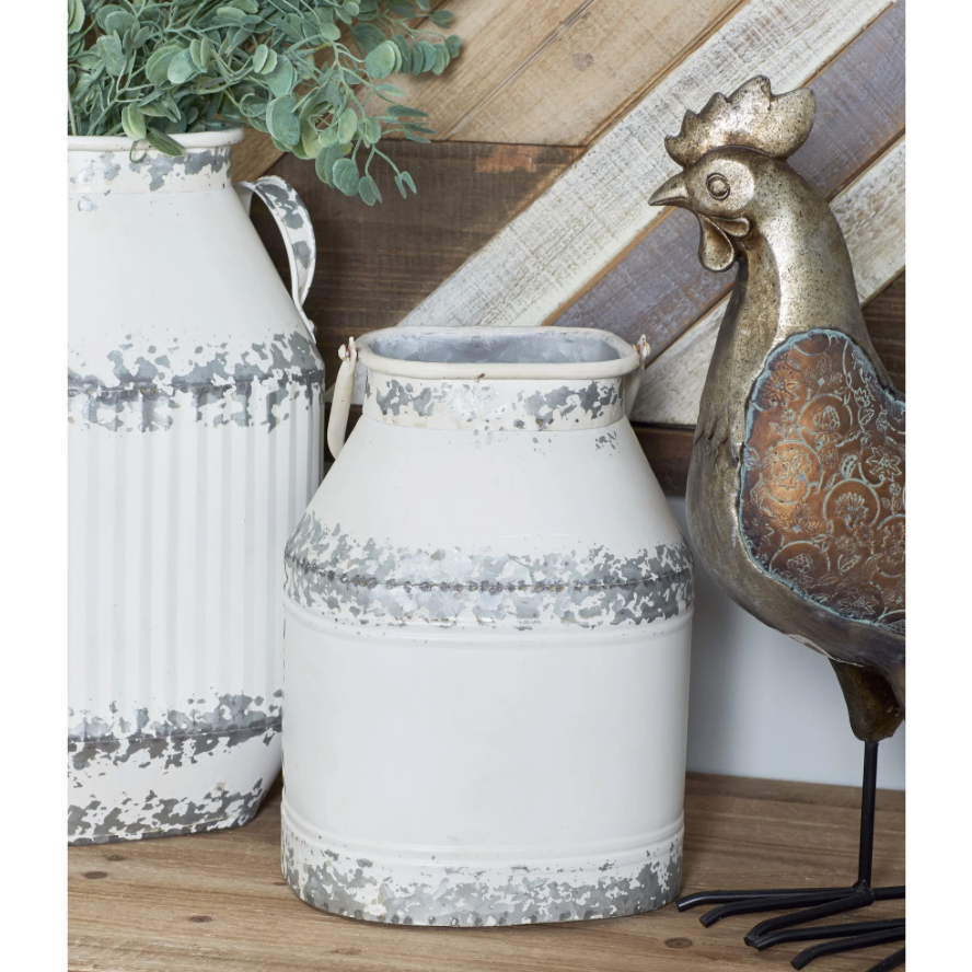 A distressed white metal milk canister vase with gray speckled bands, styled on a wooden surface next to a white ribbed vase with greenery and a decorative galvanized metal rooster figurine.