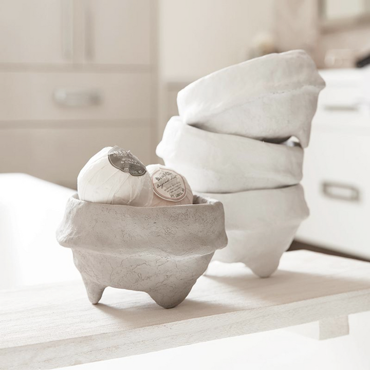 A white paper maché footed bowl with a textured surface, displayed on a wooden surface alongside a stack of similar bowls in the background.
