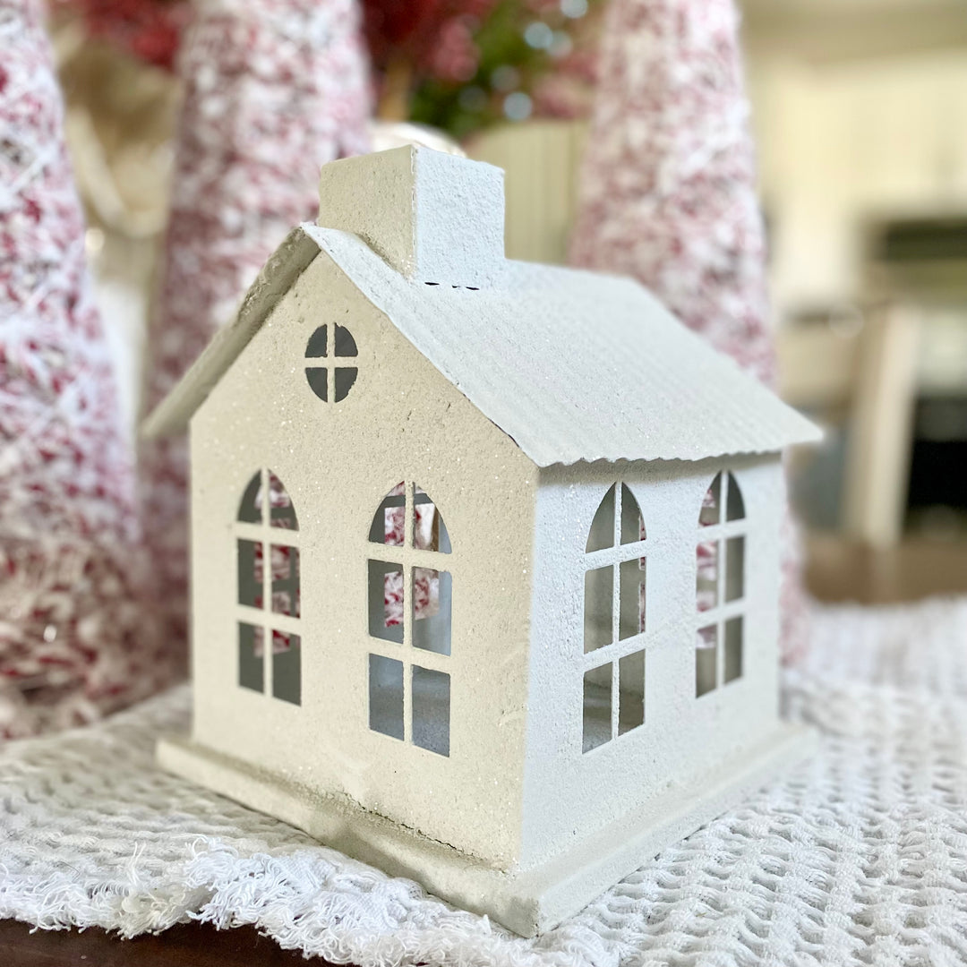 A white, metal Snowy Chalet with a textural snowy finish, topped with sparkling glitter, placed on a white textile surface.