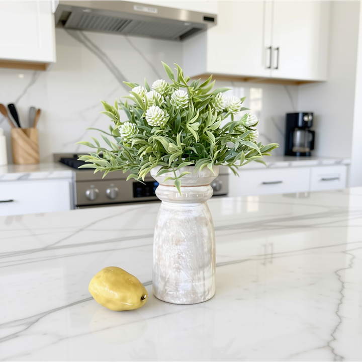 A decorative faux flower arrangement in a distressed white ceramic vase, styled on a white marble kitchen countertop with a yellow pear nearby and a blurred modern kitchen in the background.