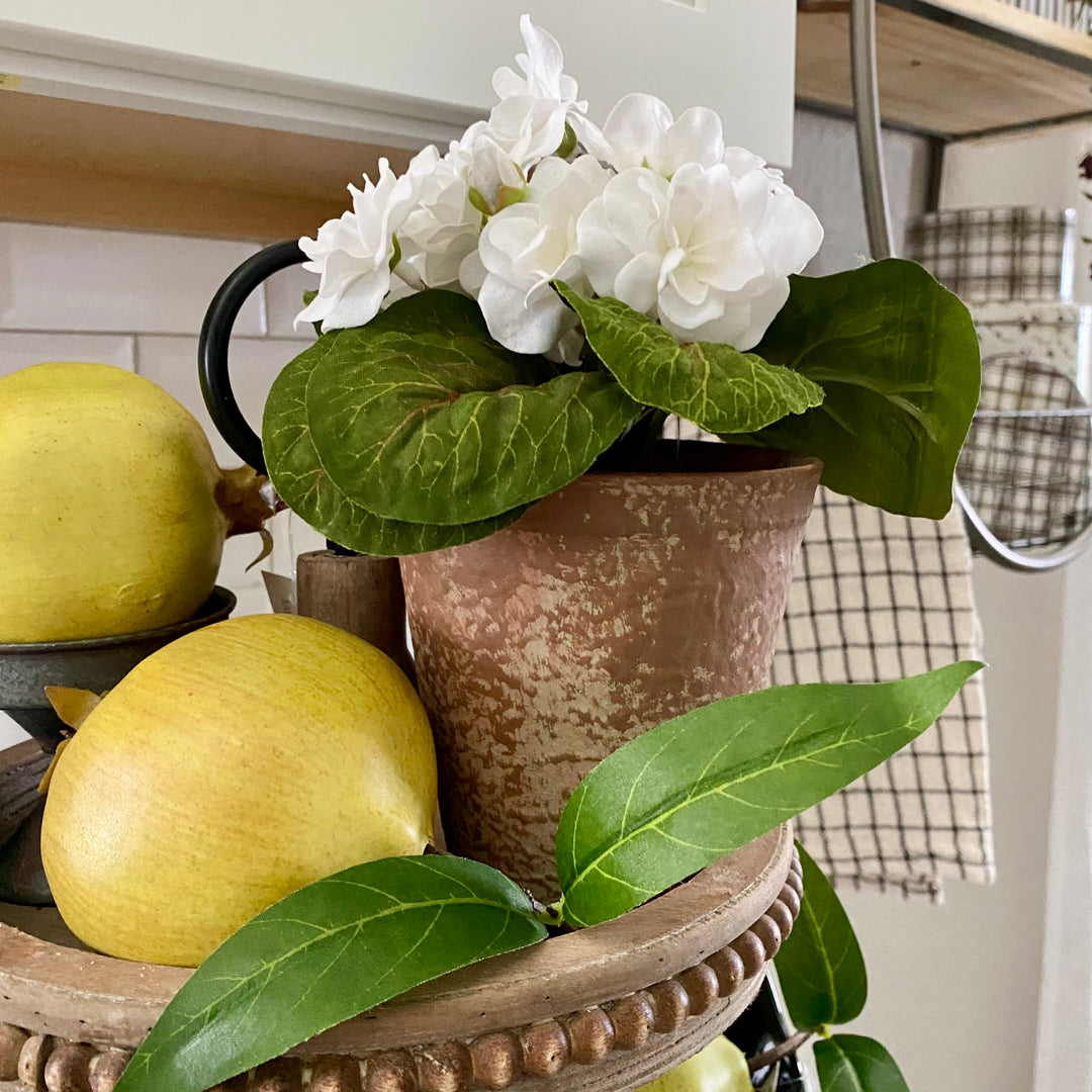 An artificial White Violet plant with green leaves in a distressed clay pot, placed on a decorative tray with faux pomegranates.