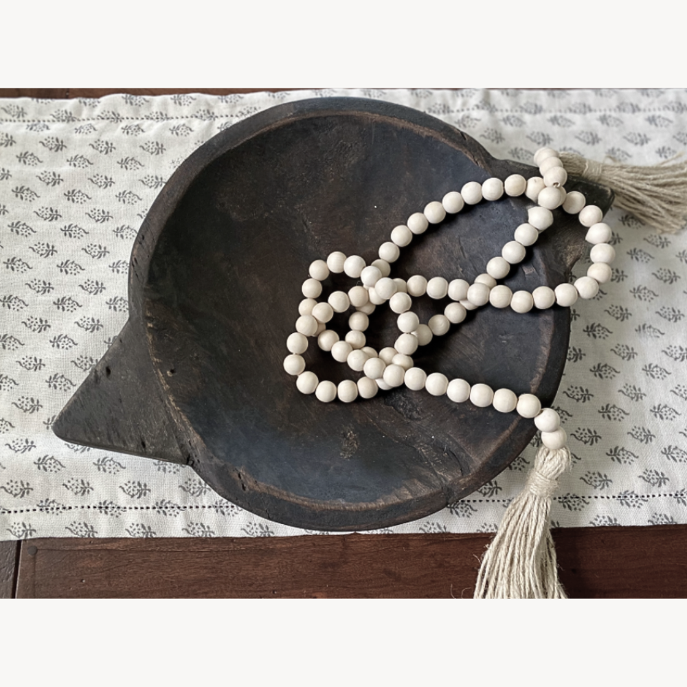 White bead garland with tassel on a dark wooden bowl on a patterned fabric table runner.