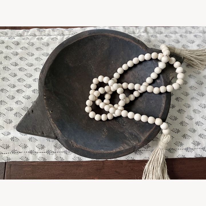 White bead garland with tassel on a dark wooden bowl on a patterned fabric table runner.