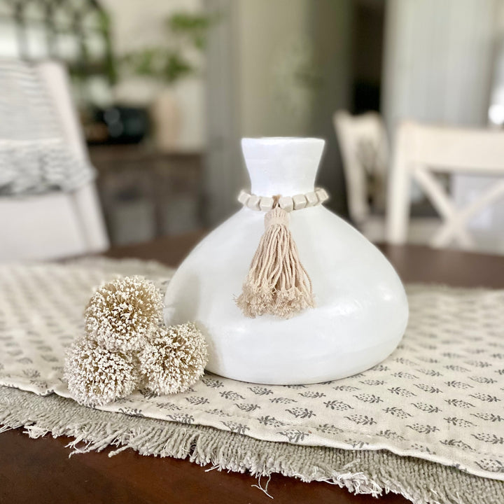 A white wood vase with ceramic beads and a tassel finish, displayed on a table with a neutral-colored tablecloth in the background.