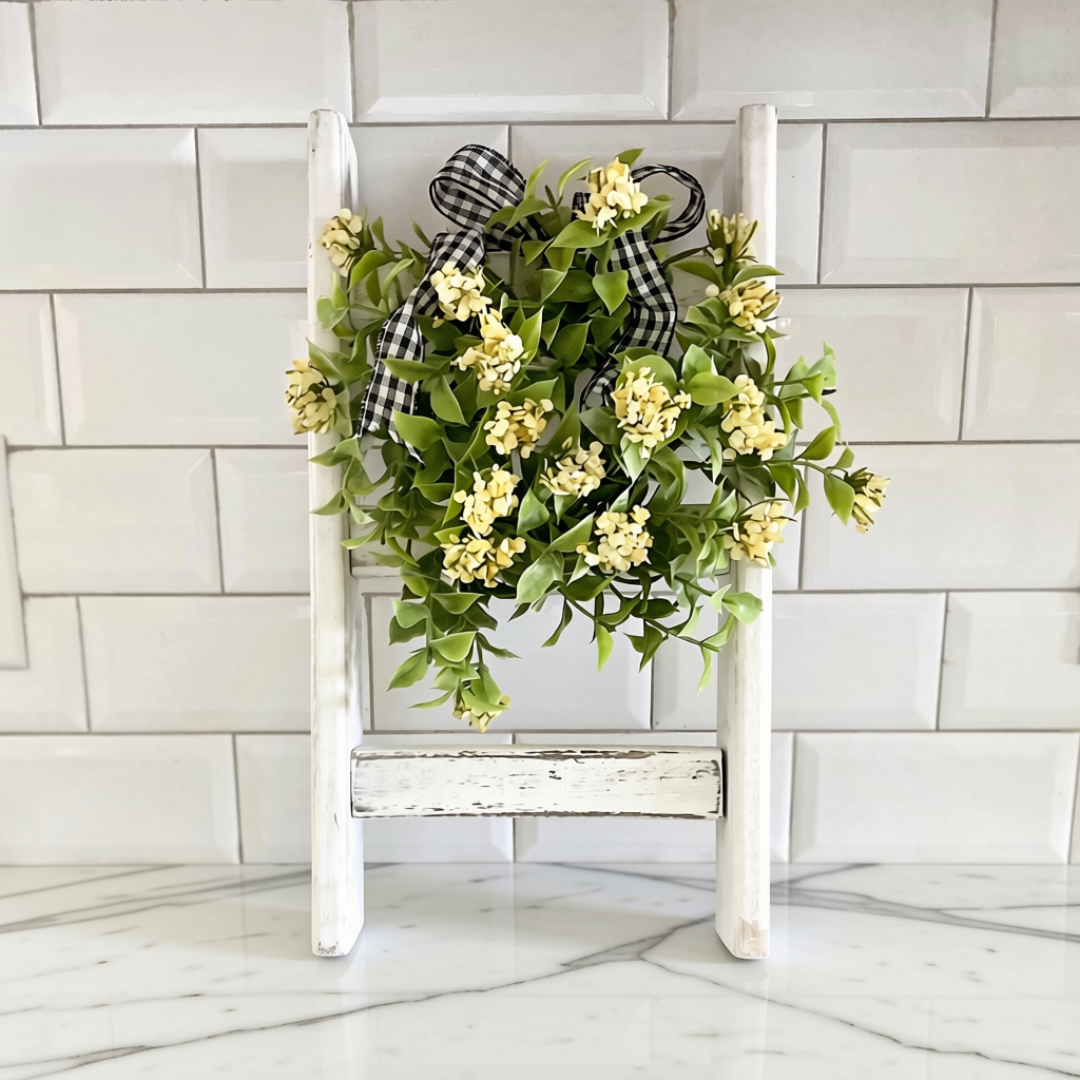 A decorative faux floral wreath with small yellow flowers, green leaves, and a black-and-white gingham ribbon bow, displayed on a white wooden countertop ladder stand against a white subway tile wall and marble countertop.