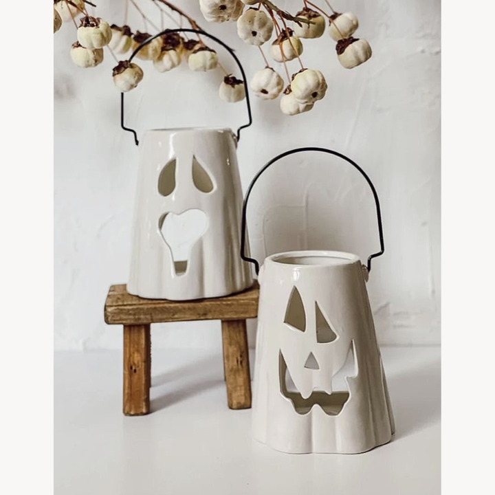 White ceramic ghost and pumpkin lanterns on a wooden stand with white berries in the background.