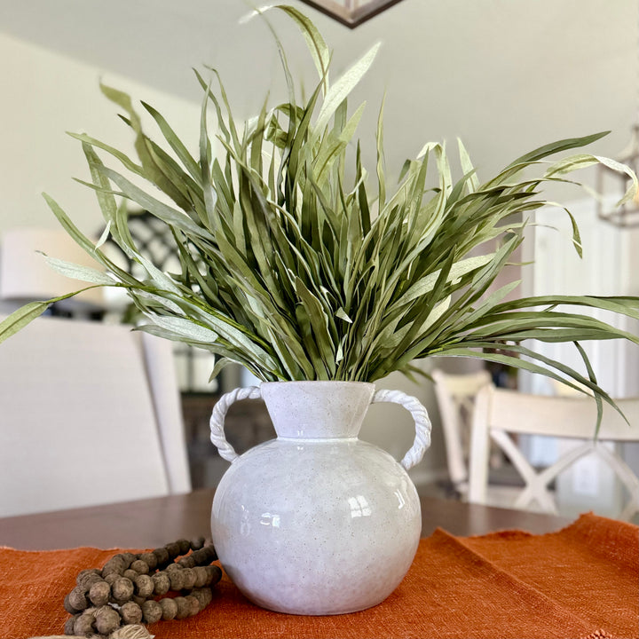 White vase with green branches, wooden beads, and small pumpkins on an orange table runner.