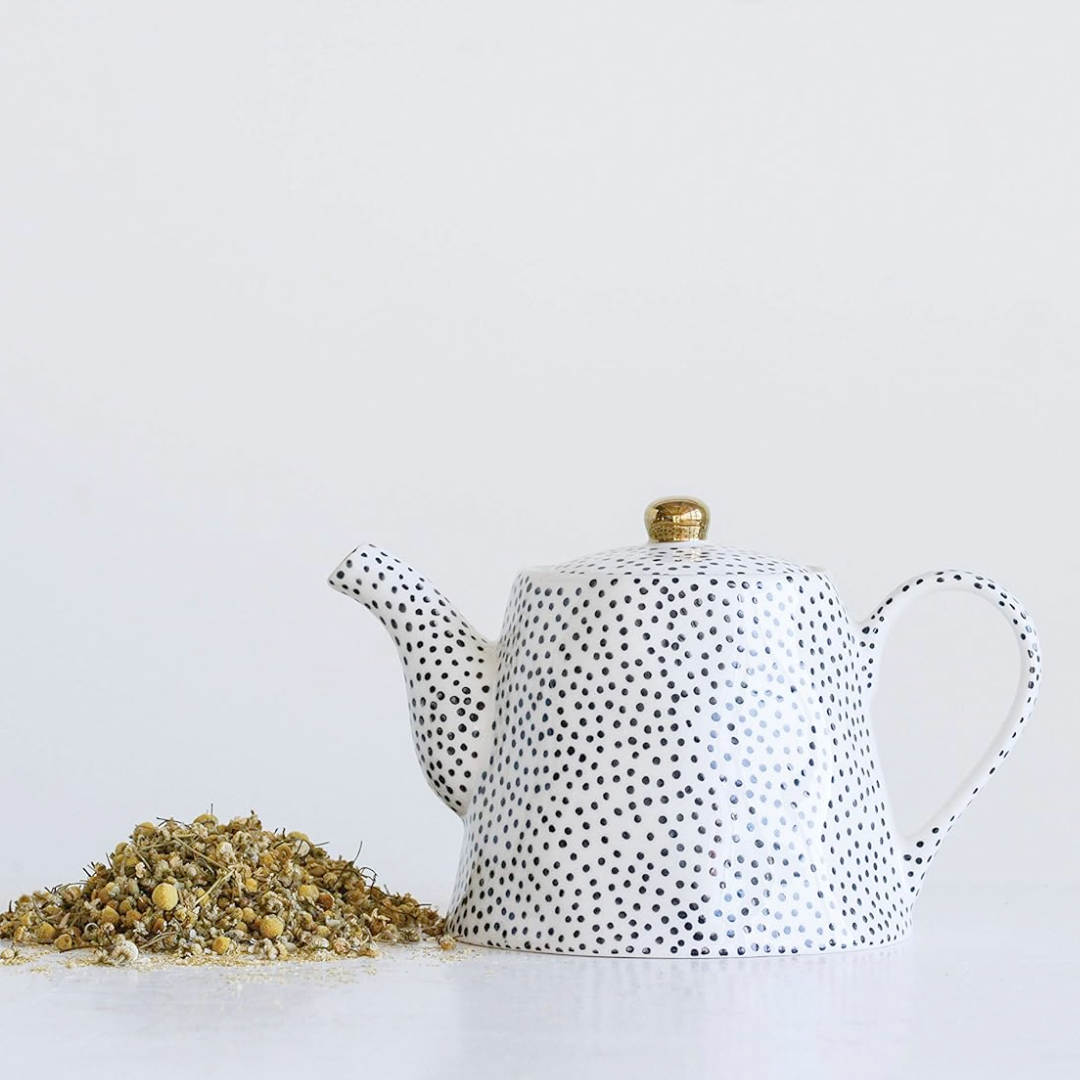 A black and white stoneware teapot with a gold-colored lid finial, displayed alongside loose tea leaves.