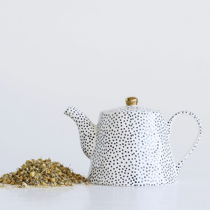 A black and white stoneware teapot with a gold-colored lid finial, displayed alongside loose tea leaves.