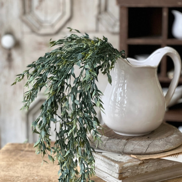 A white stoneware pitcher with a scalloped rim and side handle, displaying a decorative green plant spills over the side, set against a rustic background.