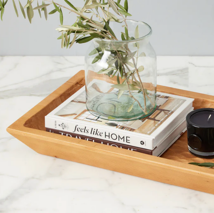 A clear glass mason jar filled with greenery, placed on top of a book titled 'Feels Like Home', inside a wooden tray on a marble surface.