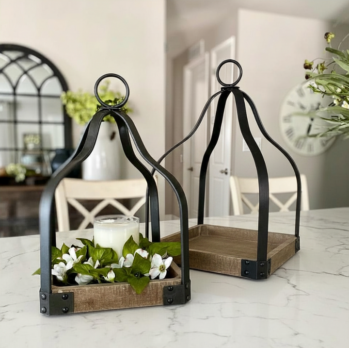 Two black metal decorative lantern-style trays with rectangular wood bases are displayed on a white marble table in a bright dining room. One tray holds white flowers and a small candle, while the other is empty.