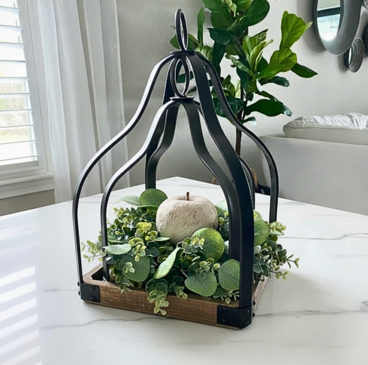 A decorative black metal lantern-style frame with a rustic wooden base, filled with green eucalyptus foliage and a small white decorative apple, set on a white marble table in a bright living room.