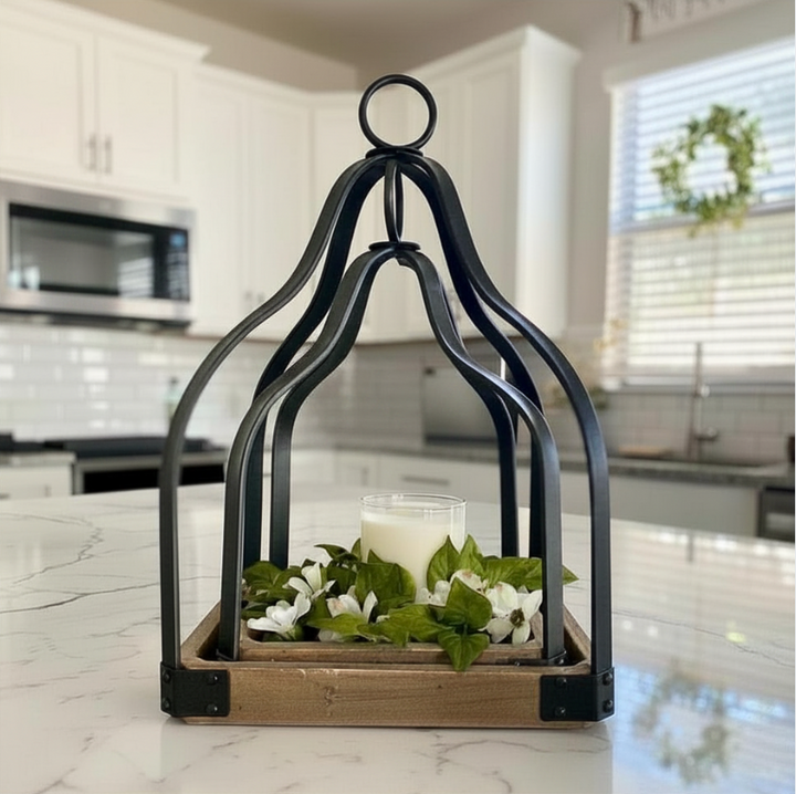 A black metal birdcage-style lantern with a rustic wooden base sits on a white marble kitchen counter, holding a white candle and green leaves with small white flowers.