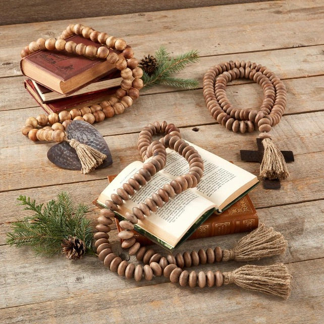 Wooden bead garlands with copper-finished pendants and jute tassels, displayed on a wooden surface with books and pine branches.