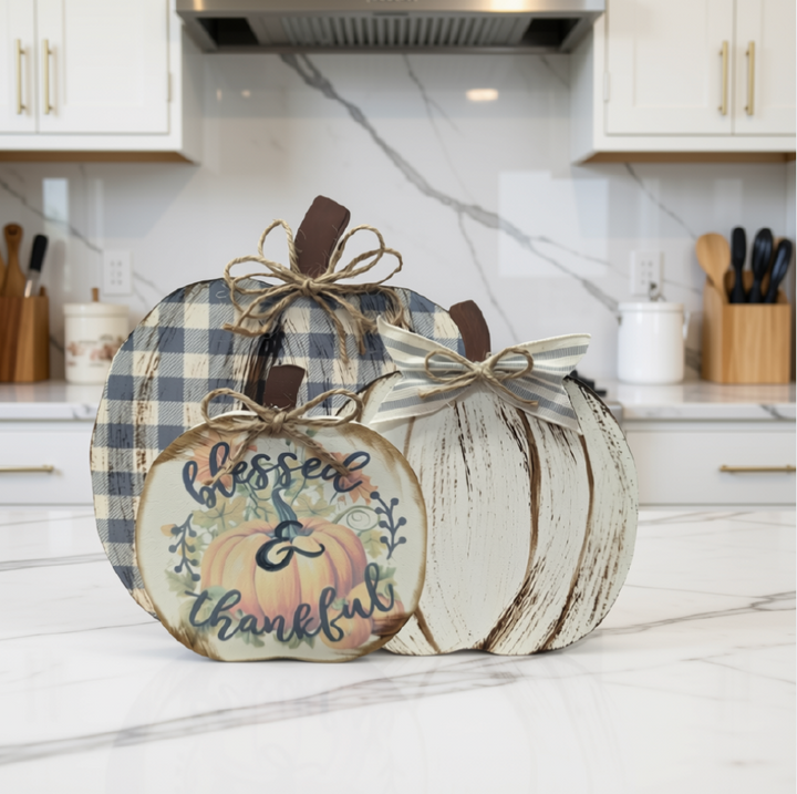 Three decorative autumn pumpkins arranged on a white marble kitchen countertop. The front pumpkin is round and painted with a warm beige and orange design featuring the words 'blessed & thankful' in script.