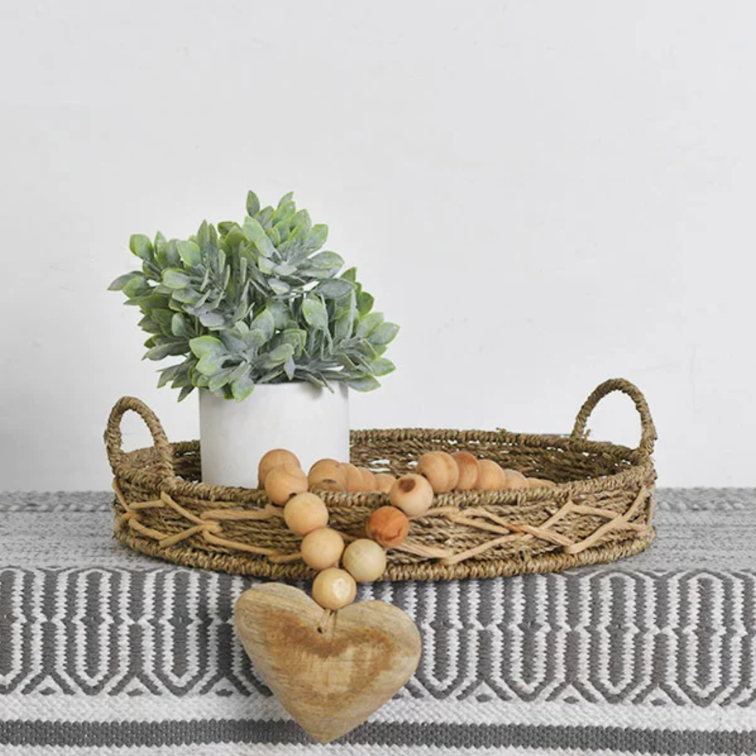 A wooden heart bead garland displayed on a woven basket, featuring a heart-shaped charm and round wooden beads. A potted plant and decorative eggs are also visible in the background.