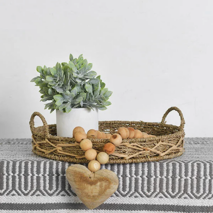 A wooden heart bead garland displayed on a woven basket, featuring a heart-shaped charm and round wooden beads. A potted plant and decorative eggs are also visible in the background.