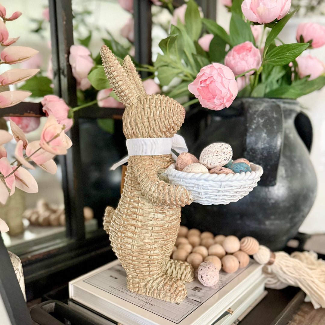 Decorative woven bunny holding a bowl of Easter eggs with pink flowers in the background.