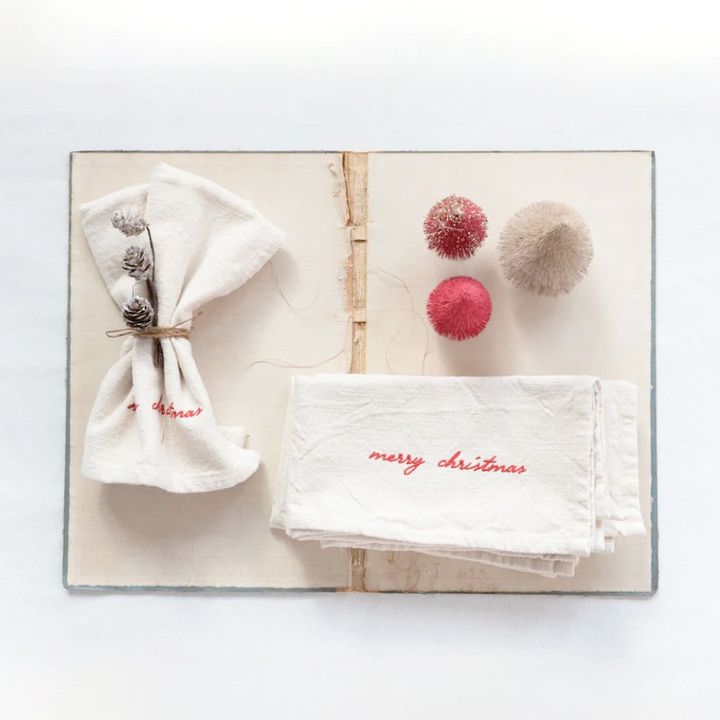 A set of four cream-colored cotton Christmas napkins with 'Merry Christmas' embroidered in red, displayed on a wooden surface with a matching table runner.