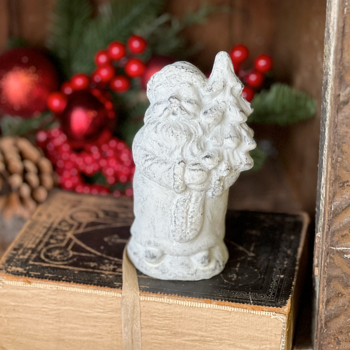 A gray concrete figurine of Santa Claus with a beard and mustache, holding a Christmas tree, placed on a box with Christmas decorations in the background.
