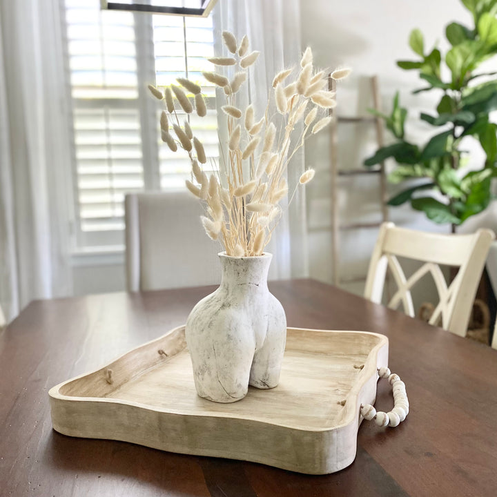 A bundle of lagrus grass in a creamy white color arranged in a wooden vase, placed on a matching wooden tray, with a neutral background featuring chairs and a plant.