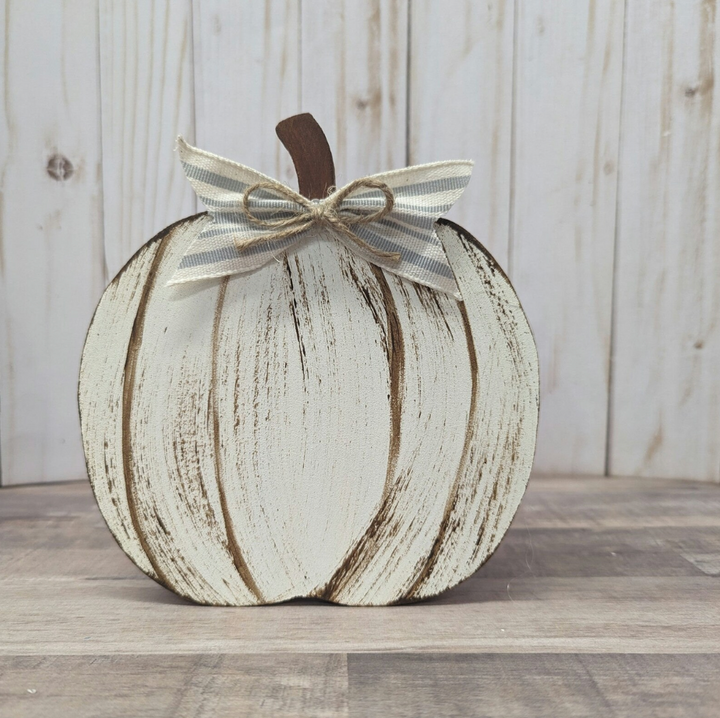 A distressed white pumpkin-shaped decorative sign with a brown stem and a striped fabric bow, displayed on a wooden surface against a whitewashed wood plank background.