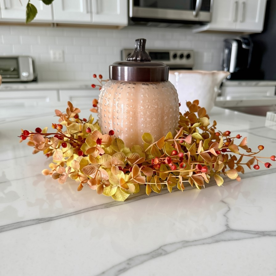 A textured glass jar candle with a dark lid, surrounded by a fall-themed faux floral wreath with yellow, orange, and red leaves, displayed on a white marble kitchen countertop.