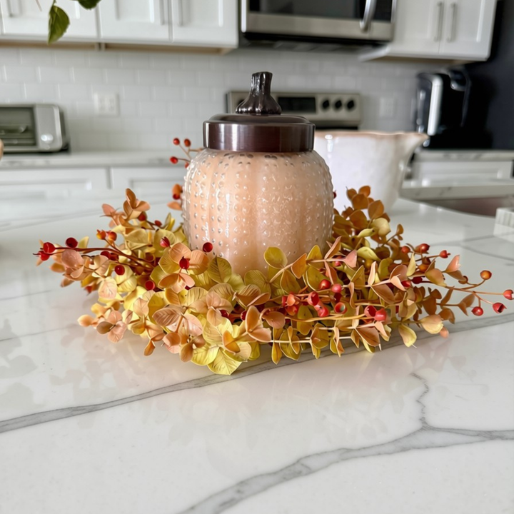 A textured glass jar candle with a dark lid, surrounded by a fall-themed faux floral wreath with yellow, orange, and red leaves, displayed on a white marble kitchen countertop.