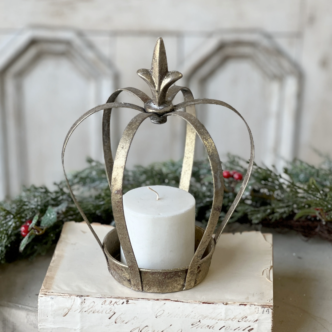 Decorative metal crown candle holder with a white candle on a wooden surface with greenery in the background.