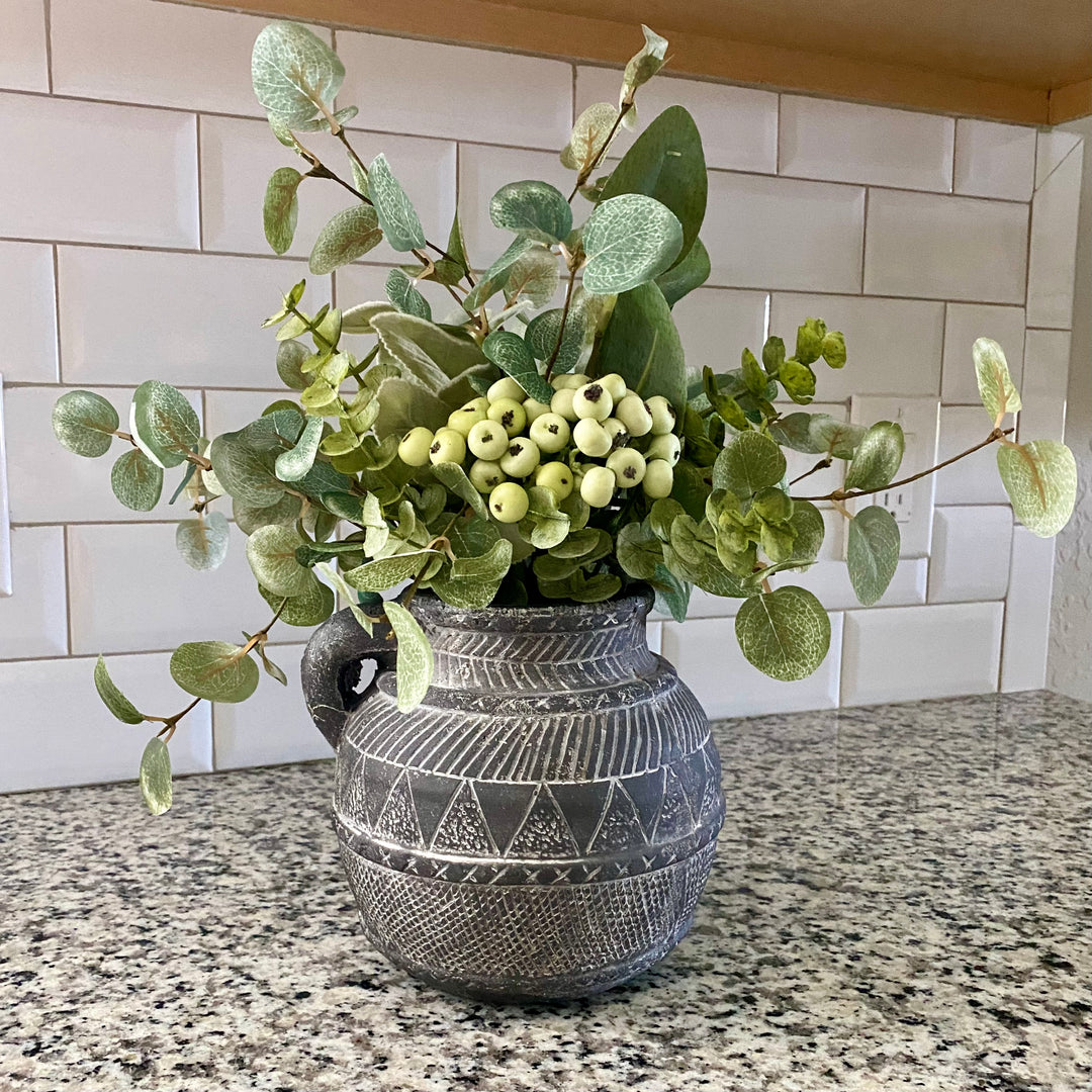 A gray terracotta vase with an aztec etched pattern filled with artificial greenery, placed on a kitchen counter.