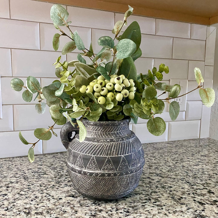 A gray terracotta vase with an aztec etched pattern filled with artificial greenery, placed on a kitchen counter.